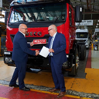 Two men in business attire shake hands in front of a red Iveco truck in a manufacturing facility. The setting features industrial elements such as machinery and safety signage, emphasizing a professional atmosphere. One man holds a document, indicating a formal agreement or partnership.