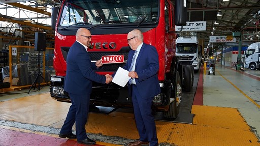 Two men in business attire shake hands in front of a red Iveco truck in a manufacturing facility. The setting features industrial elements such as machinery and safety signage, emphasizing a professional atmosphere. One man holds a document, indicating a formal agreement or partnership.