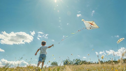 A young boy flying a kite in a sunny, open field with wildflowers under a bright blue sky with scattered clouds. The sun shines brightly above, creating a warm and cheerful atmosphere.