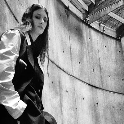 Black and white photo of a person with long hair wearing a two-tone jacket, leaning against a curved concrete wall with visible bolts, next to a metal staircase under a slatted roof.