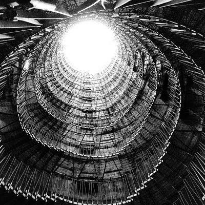 Black and white photo of a spiral staircase taken from the bottom looking up towards a bright circular opening at the top, showcasing intricate metal railings and geometric patterns.