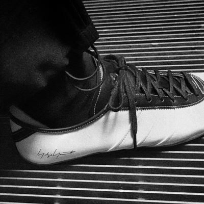 A close-up black and white photo of a person's foot wearing a stylish sneaker with laces, resting on a striped surface. The sneaker has a signature or logo on the side near the heel.