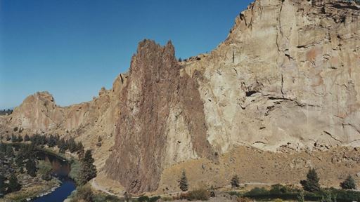 A scenic landscape featuring towering rocky cliffs with a prominent dark brown rock formation in the center, surrounded by dry vegetation and scattered trees. A winding river flows along the base of the cliffs under a clear blue sky.
