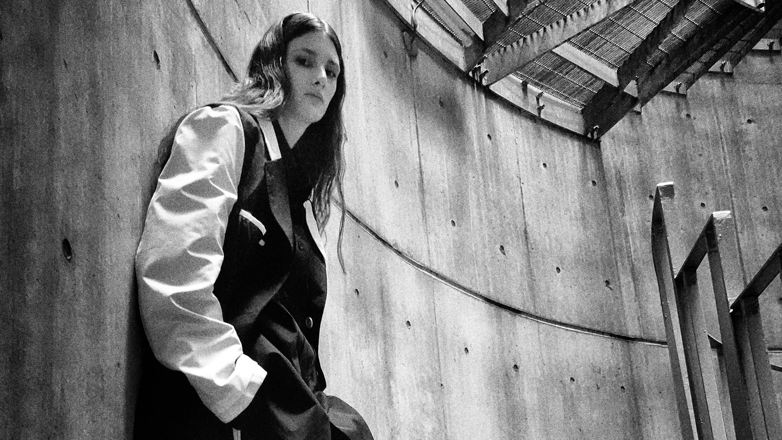 Black and white photo of a person with long hair wearing a two-tone jacket, leaning against a curved concrete wall with visible bolts, next to a metal staircase under a slatted roof.