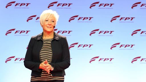 A woman with short blonde hair stands on stage in front of a backdrop featuring the FPT logo. She is wearing a black jacket over a striped shirt and appears to be speaking or presenting. The background is a light blue with repeated FPT branding.